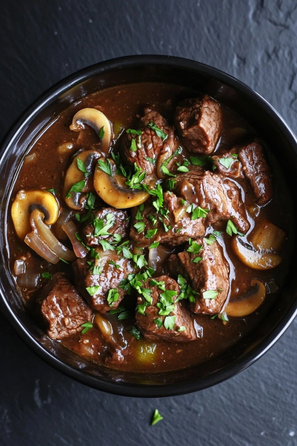 Crock pot beef marsala - the image is a close-up of a dish in a black bowl. the dish appears to be a beef stew with chunks of beef, mushrooms, and onions. the beef is cooked in a rich, dark brown sauce and is garnished with chopped parsley. the mushrooms are sliced and yellow, and the onions are caramelized. the sauce is thick and thick, and there are small pieces of herbs scattered throughout the dish. the bowl is sitting on a dark, textured surface.