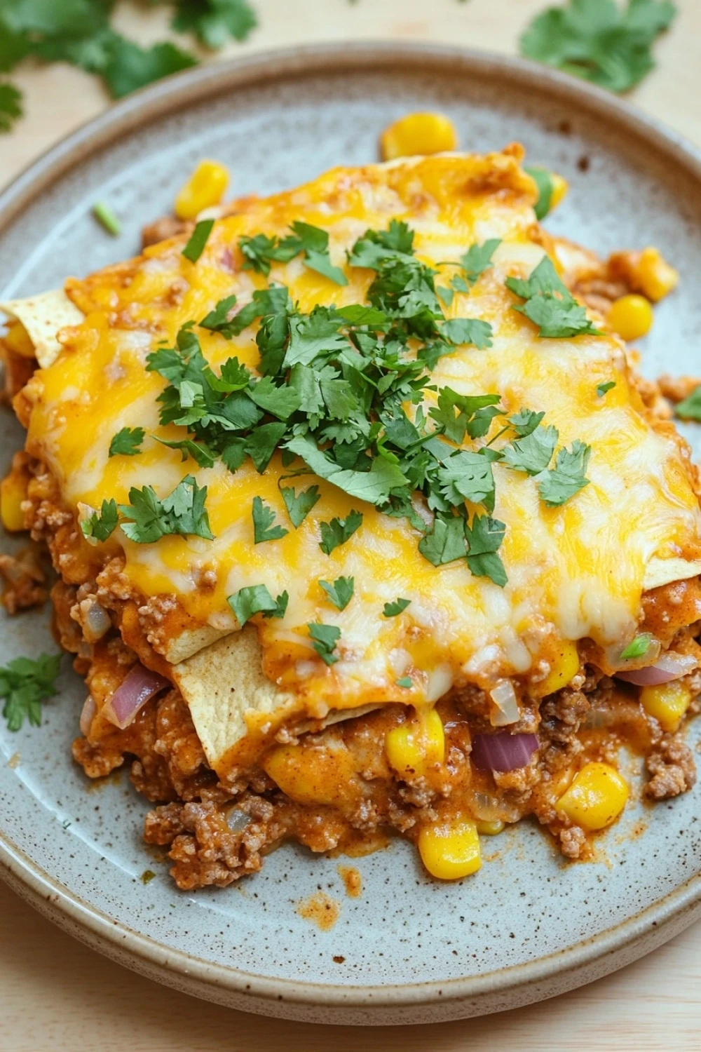 Crockpot ground beef enchilada casserole - the image shows a plate of enchiladas on a wooden table. the plate is light blue with a speckled pattern and is garnished with fresh cilantro. the enchillas are golden brown and appear to be made with ground beef, corn kernels, and red onions. they are arranged in a pile on top of each other and are covered in melted cheese. there are also a few sprigs of cilantro scattered around the plate. the background is blurred, but it appears to be a kitchen countertop.
