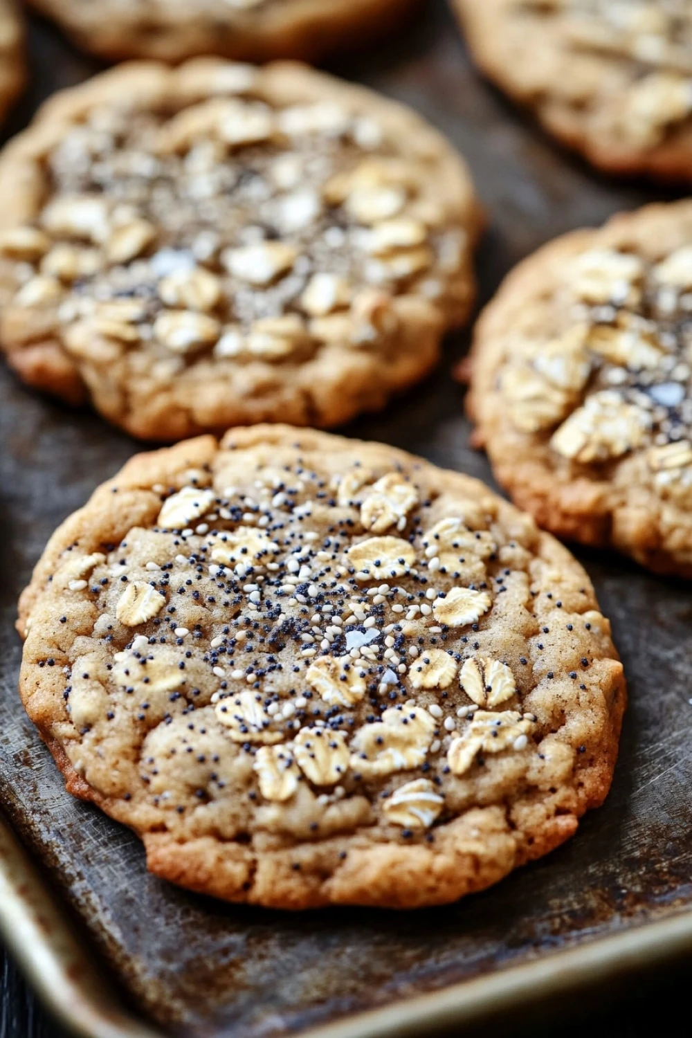 Everything oatmeal cookie - the image shows a baking tray lined with freshly baked cookies. the cookies are round and golden brown in color, with visible chunks of nuts and seeds scattered throughout. the tray is lined with parchment paper, and the cookies are arranged in a neat row. the background is blurred, but it appears to be a wooden table or countertop.