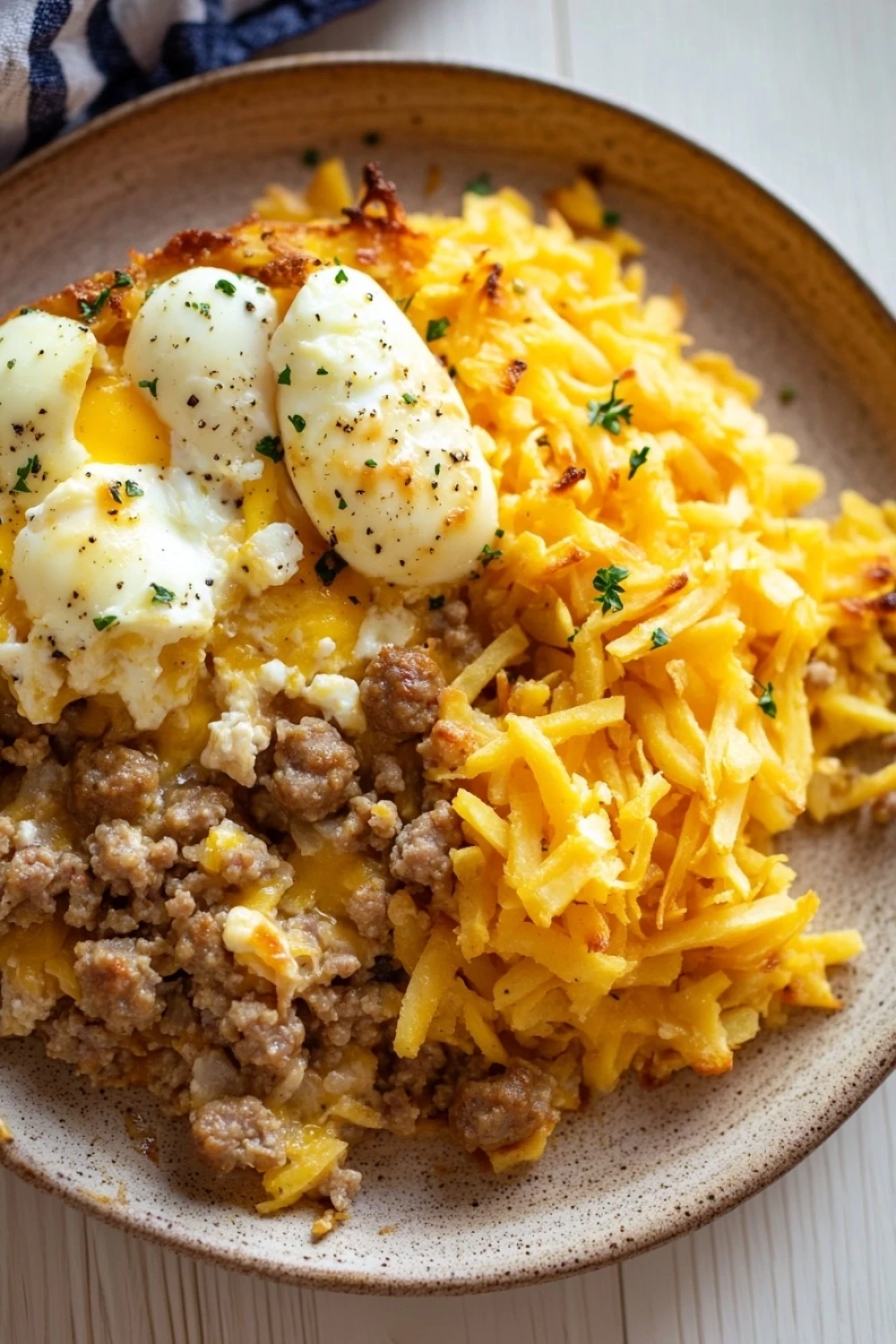 Freezer breakfast casserole hashbrown - the image is a close-up of a plate of food. the plate is made of ceramic and has a brown rim. on top of the plate, there is a serving of scrambled eggs with a poached egg on top. the eggs are cooked sunny-side up and have a runny yolk. the scrambled eggs are yellow and appear to be seasoned with herbs and spices. the dish is served on a white wooden table with a blue and white striped napkin in the background.