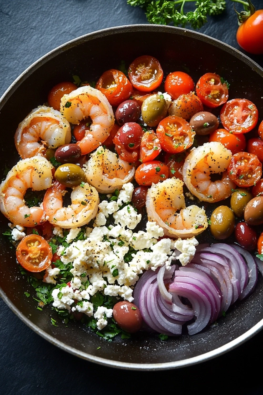 Greek shrimp and feta skillet - the image is a close-up of a salad in a black frying pan. the salad is made up of various ingredients, including shrimp, cherry tomatoes, olives, red onions, and feta cheese. the shrimp are arranged in a circular pattern, with some overlapping each other. the cherry tomatoes are bright red and appear to be ripe, while the olives are dark green. the feta is crumbled and scattered throughout the salad. there are also a few sprigs of parsley scattered around the pan, adding a pop of green color to the dish. the pan is sitting on a dark grey countertop.