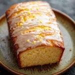 Greek yogurt lemon loaf cake - the image shows a slice of cake on a brown plate. the cake appears to be freshly baked and has a golden brown crust on top. it is drizzled with a white glaze that is dripping down the sides of the cake. the glaze is decorated with small yellow flowers, adding a pop of color to the dish. the plate is sitting on a dark, textured surface, possibly a table or countertop.