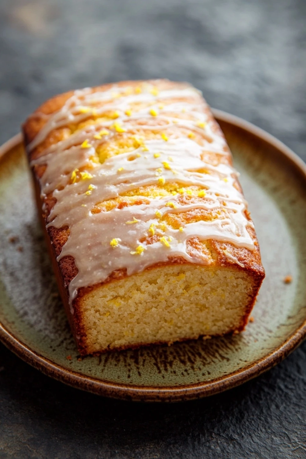 Greek yogurt lemon loaf cake - the image shows a slice of cake on a brown plate. the cake appears to be freshly baked and has a golden brown crust on top. it is drizzled with a white glaze that is dripping down the sides of the cake. the glaze is decorated with small yellow flowers, adding a pop of color to the dish. the plate is sitting on a dark, textured surface, possibly a table or countertop.