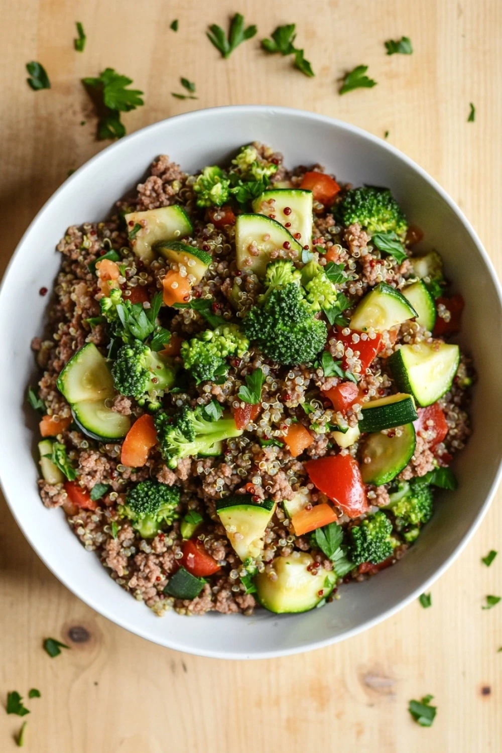 Ground beef dinner macro friendly - the image is a close-up of a bowl of quinoa salad. the bowl is white and is placed on a wooden table. the quinoa is a light brown color and is mixed with various vegetables such as broccoli, zucchini, carrots, and bell peppers. the vegetables are bright green and appear to be seasoned with herbs and spices. there are also a few sprigs of parsley scattered around the bowl. the overall appearance of the salad is colorful and appetizing.