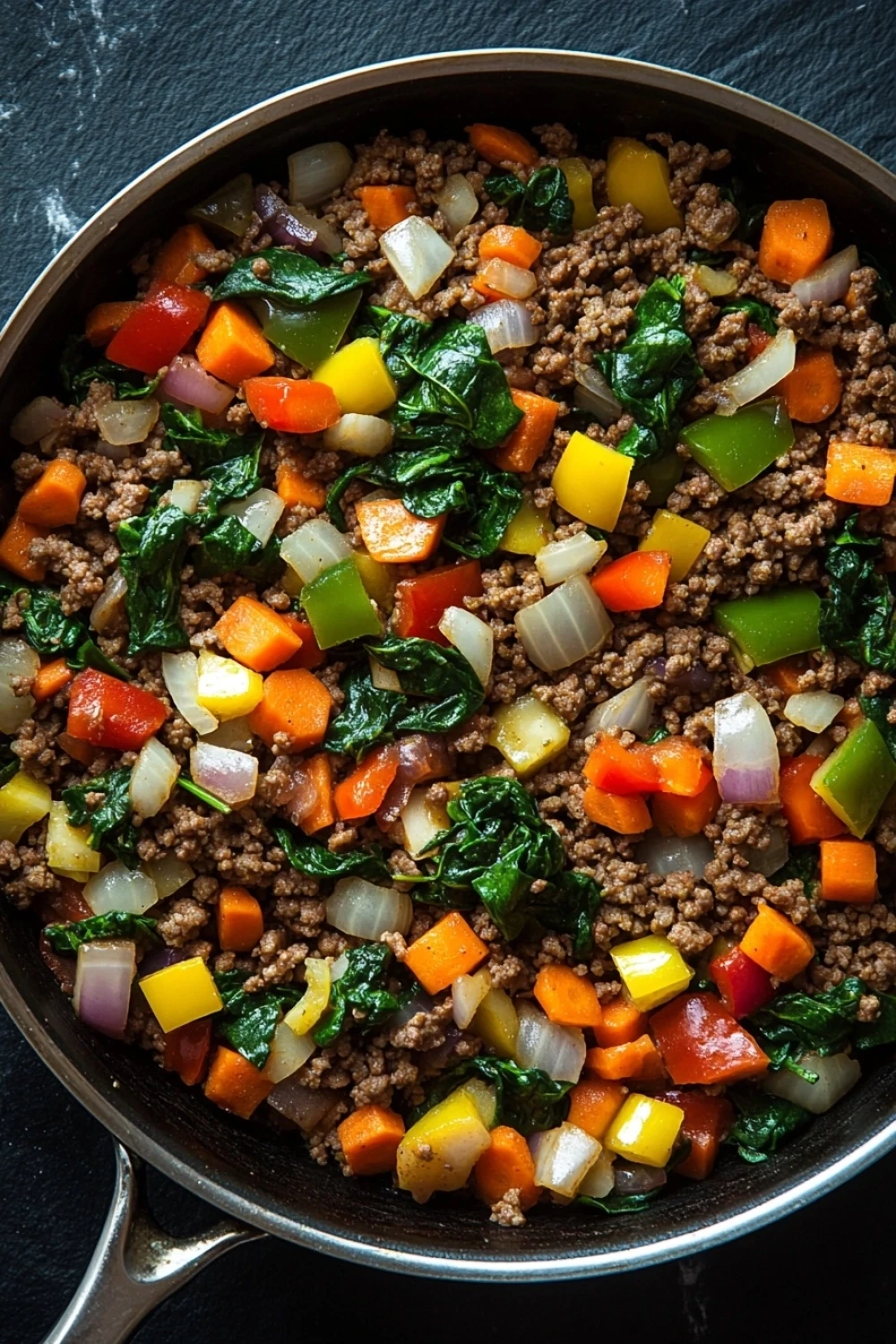 Ground beef dinner no pasta - the image shows a frying pan with a mixture of ground beef and vegetables in it. the pan is black and has a handle on one side. the ground beef appears to be cooked and is mixed with various vegetables such as carrots, bell peppers, onions, and spinach. the vegetables are bright orange and yellow in color and are arranged in a colorful and appetizing manner. the background is dark and out of focus, making the colors of the vegetables stand out.