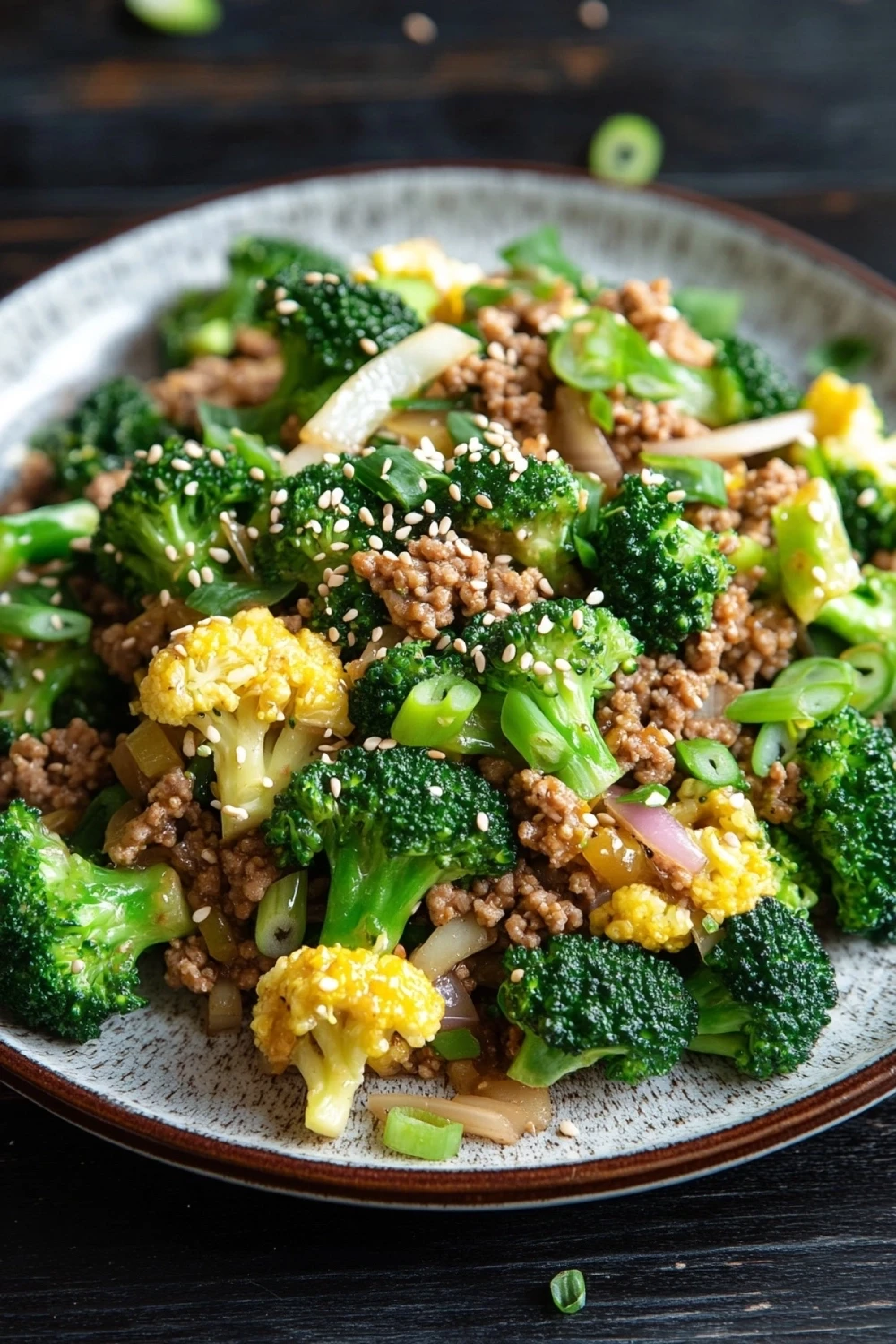 Ground turkey and broccoli for dinner - the image is a close-up of a plate of food. the plate is white with a brown rim and is placed on a dark wooden table. the food appears to be a stir-fry dish with broccoli, cauliflower, and minced meat. the broccoli is bright green and looks fresh, while the cauliflower is yellow and fluffy. the minced meat is a light brown color and is scattered throughout the dish. there are also some sesame seeds sprinkled on top. the dish is garnished with sliced onions and green onions.
