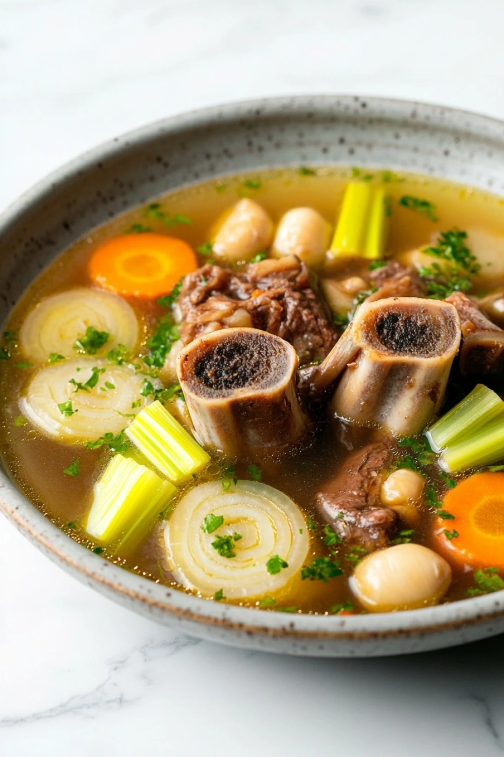 High protein beef bone broth - the image is a close-up of a bowl of soup. the soup appears to be thick and creamy, with chunks of meat and vegetables mixed in. the broth is a light brown color and is thick and thick. there are also chunks of carrots, celery, and onions visible in the soup. on top of the soup, there is a sprig of parsley. the bowl is made of ceramic and is sitting on a white marble countertop.