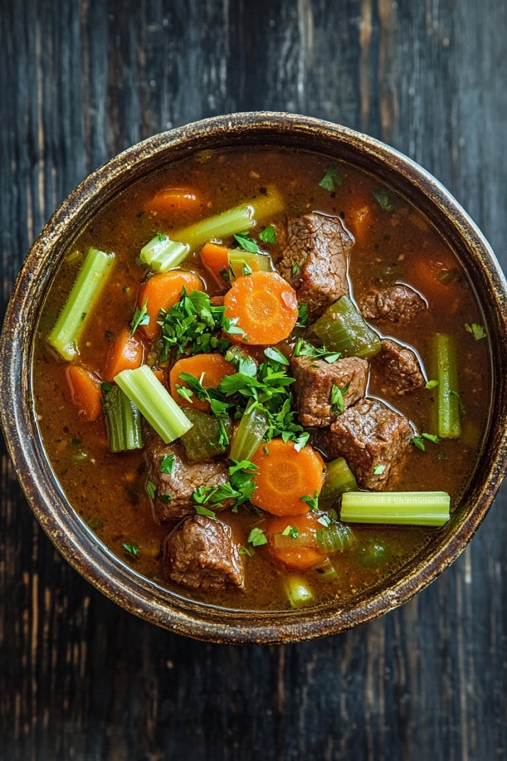 High protein beef crockpot - the image is a close-up of a bowl of beef stew on a dark wooden table. the stew is made with chunks of beef, carrots, celery, and herbs, and is garnished with fresh parsley. the bowl is made of ceramic and has a dark brown color. the background is blurred, but it appears to be a rustic wooden surface. the overall mood of the image is warm and inviting.