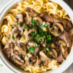 High protein beef stroganoff crockpot - the image shows a bowl of beef stroganoff with fettuccine noodles. the beef is cooked medium-rare and is covered in a creamy mushroom sauce. the noodles are long and thin, and they are arranged in a pile on top of each other. the dish is garnished with fresh parsley. the bowl is white and is sitting on a wooden table. the background is blurred, but it appears to be a rustic kitchen countertop.
