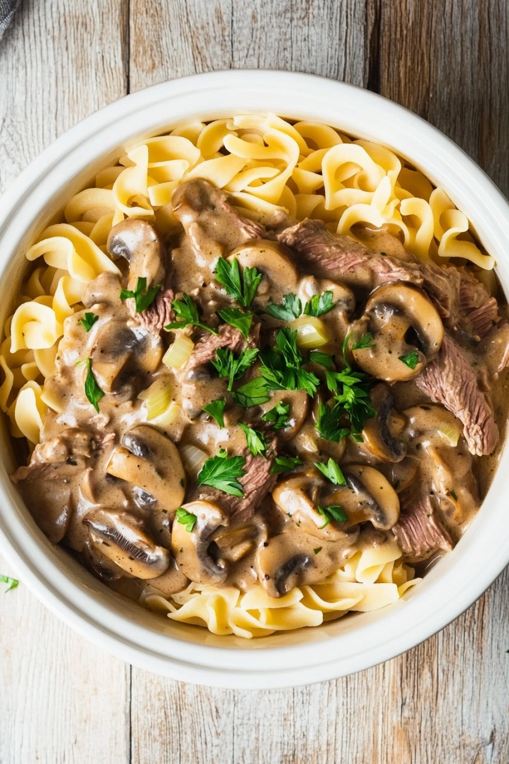 High protein beef stroganoff crockpot - the image shows a bowl of beef stroganoff with fettuccine noodles. the beef is cooked medium-rare and is covered in a creamy mushroom sauce. the noodles are long and thin, and they are arranged in a pile on top of each other. the dish is garnished with fresh parsley. the bowl is white and is sitting on a wooden table. the background is blurred, but it appears to be a rustic kitchen countertop.