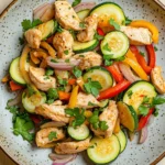 High protein chicken and zucchini - the image is a close-up of a plate of salad. the plate is round and has a light blue and white speckled pattern. the salad is made up of various vegetables, including zucchini, red bell peppers, yellow squash, and red onions. there are also chunks of chicken mixed in with the vegetables. the chicken is cooked to a golden brown color and appears to be seasoned with herbs and spices. the vegetables are arranged in a colorful and appetizing manner, with some overlapping each other. the dish is garnished with fresh cilantro leaves.