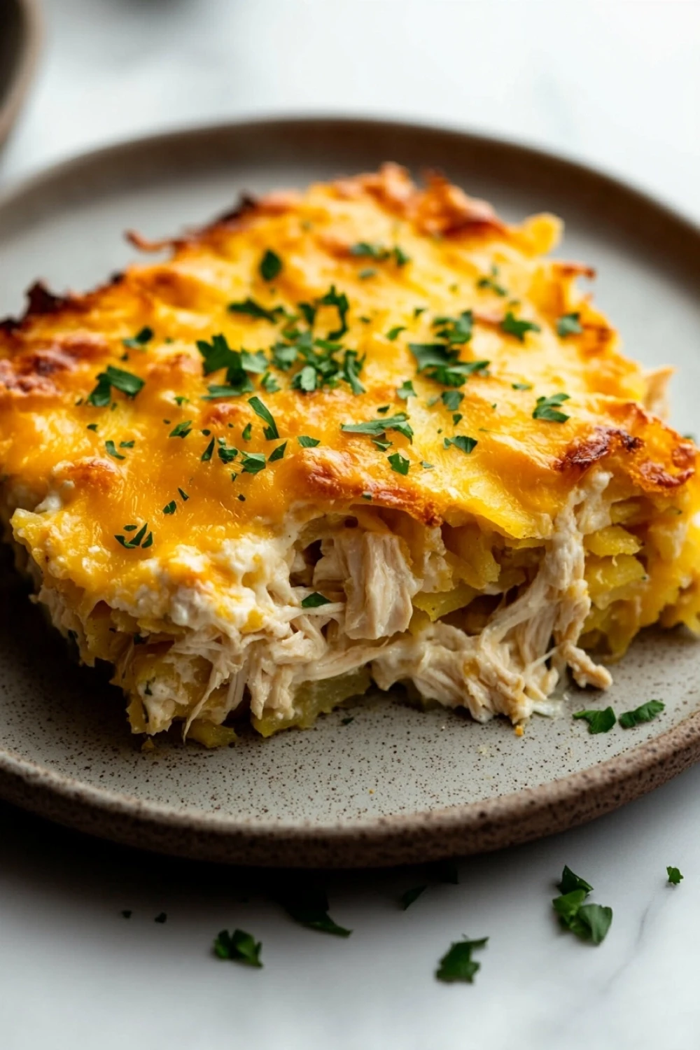 High protein chicken hashbrown casserole - the image shows a slice of chicken casserole on a brown plate. the dish is golden brown and has a layer of melted cheese on top. the cheese is melted and bubbly, and there are small pieces of chicken scattered throughout the dish. the plate is garnished with chopped parsley. the background is blurred, but it appears to be a kitchen countertop.