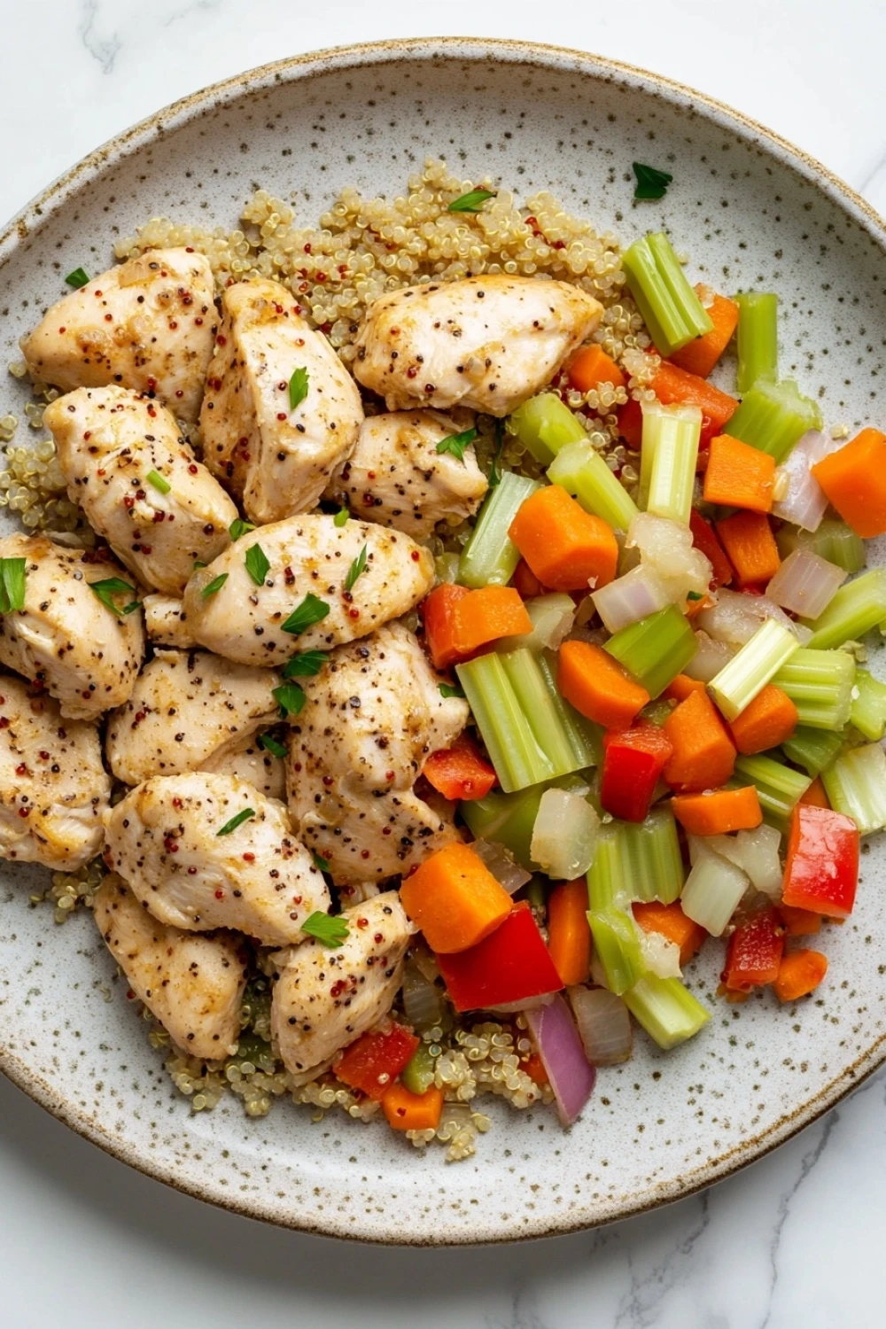 High protein chicken in crockpot - the image is a close-up of a plate of food. the plate is white with a speckled pattern and is placed on a white marble countertop. on top of the plate, there are several pieces of cooked chicken, which appear to be seasoned with herbs and spices. the chicken is cut into small cubes and is arranged in a pile on top of a bed of quinoa. on the right side of the image, there is a pile of diced carrots, celery, and red onions. the vegetables are cut into thin strips and are mixed in with the quinoa, creating a colorful and appetizing dish.
