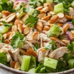 High protein chicken salad with greek yogurt - the image is a close-up of a bowl of salad. the bowl is made of ceramic and has a speckled pattern. the salad is made up of various ingredients, including shredded chicken, sliced red onions, celery, and cilantro. the chicken is cut into small pieces and appears to be seasoned with herbs and spices. there are also sliced almonds scattered throughout the salad, adding a crunchy texture. the colors of the ingredients are vibrant and the dish looks fresh and appetizing.