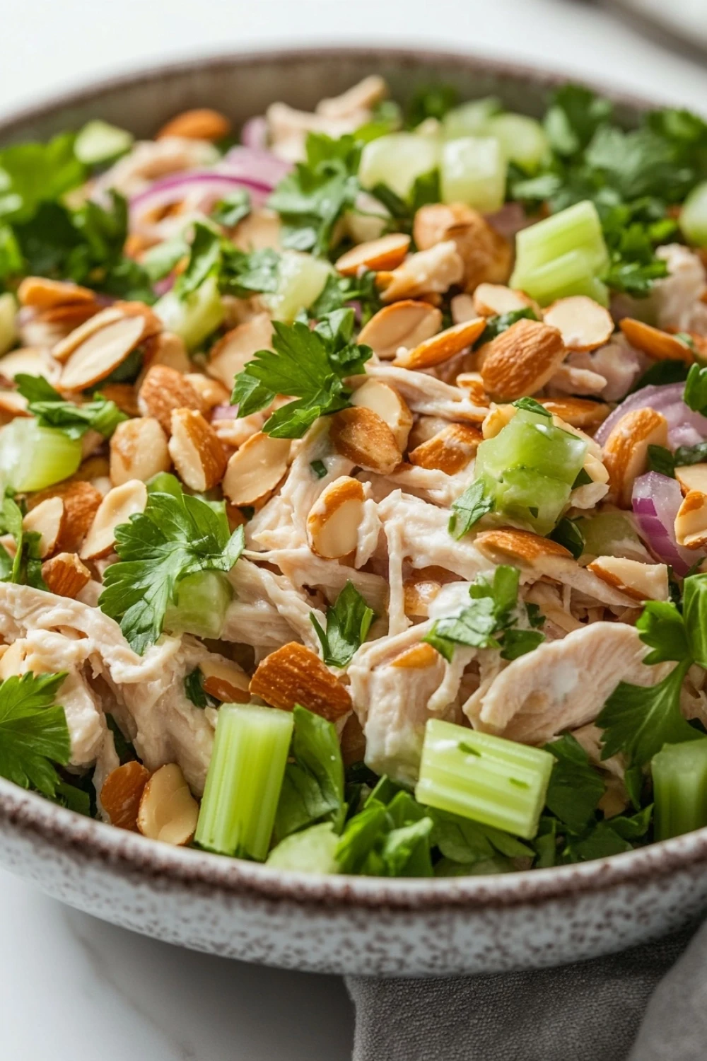 High protein chicken salad with greek yogurt - the image is a close-up of a bowl of salad. the bowl is made of ceramic and has a speckled pattern. the salad is made up of various ingredients, including shredded chicken, sliced red onions, celery, and cilantro. the chicken is cut into small pieces and appears to be seasoned with herbs and spices. there are also sliced almonds scattered throughout the salad, adding a crunchy texture. the colors of the ingredients are vibrant and the dish looks fresh and appetizing.