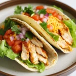 High protein chicken tacos - the image shows two tacos on a brown plate. the tacos are made with a tortilla shell and are filled with shredded chicken, diced tomatoes, red onions, and cilantro. the chicken is cooked to a golden brown color and appears to be seasoned with herbs and spices. the salad is made up of lettuce, tomatoes, and other vegetables, and there is a layer of shredded cheese on top. the plate is sitting on a white tablecloth.
