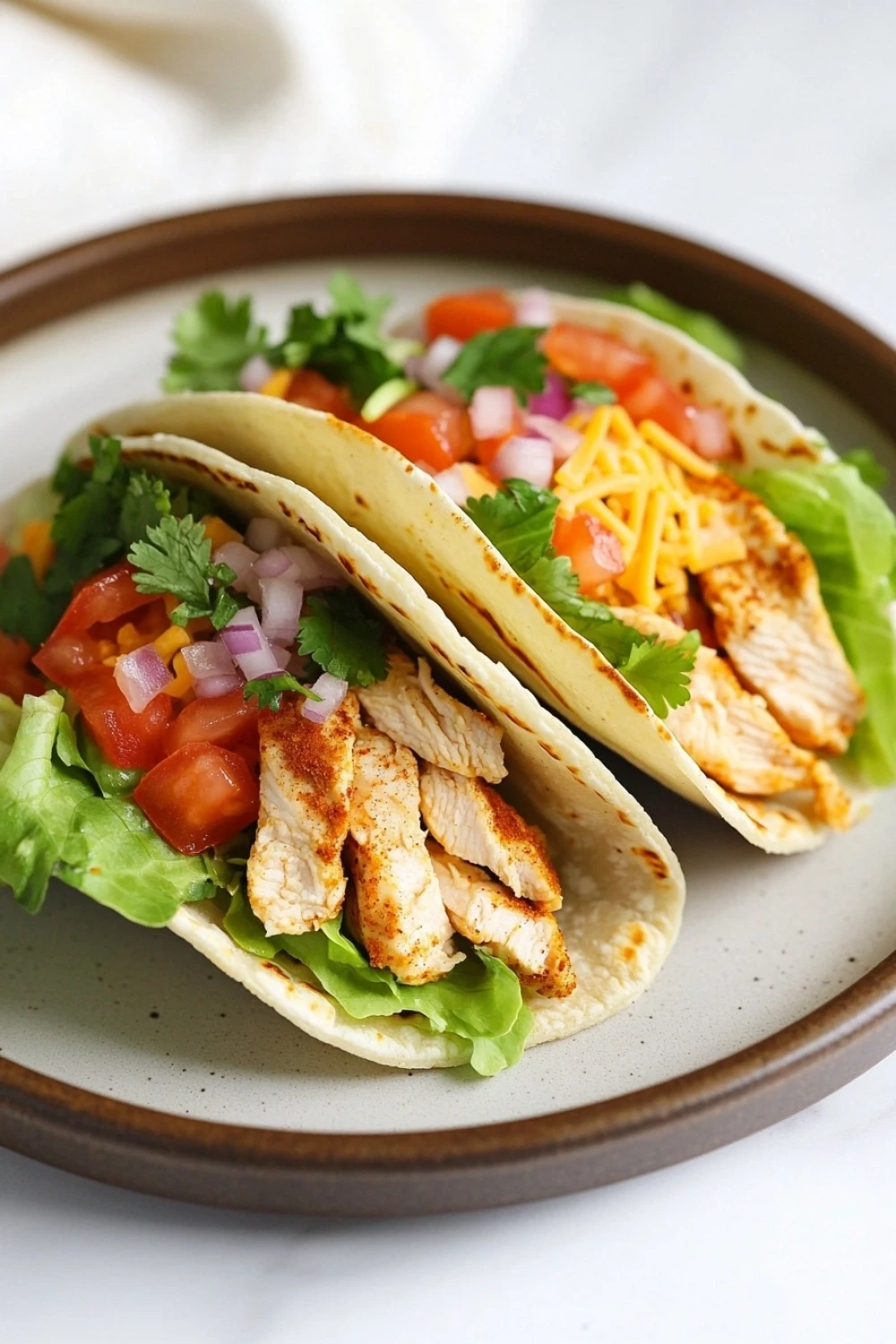 High protein chicken tacos - the image shows two tacos on a brown plate. the tacos are made with a tortilla shell and are filled with shredded chicken, diced tomatoes, red onions, and cilantro. the chicken is cooked to a golden brown color and appears to be seasoned with herbs and spices. the salad is made up of lettuce, tomatoes, and other vegetables, and there is a layer of shredded cheese on top. the plate is sitting on a white tablecloth.