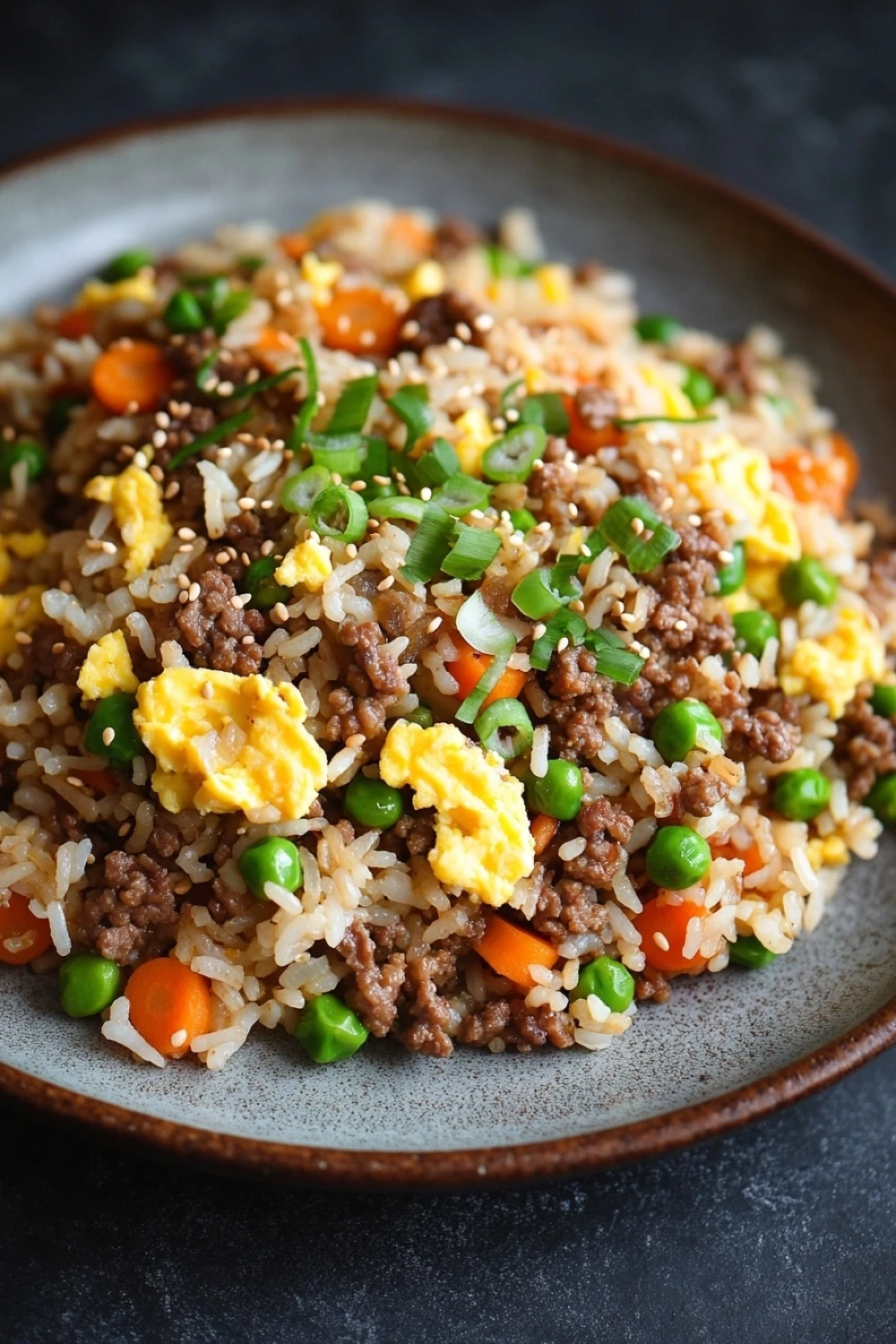 High protein korean beef fried rice - the image is a close-up of a plate of fried rice with minced meat and vegetables. the rice is brown and appears to be seasoned with sesame seeds. there are small pieces of green onions scattered throughout the dish, adding a pop of color. the dish is served on a dark grey plate with a textured surface. the background is blurred, making the plate the focal point of the image.