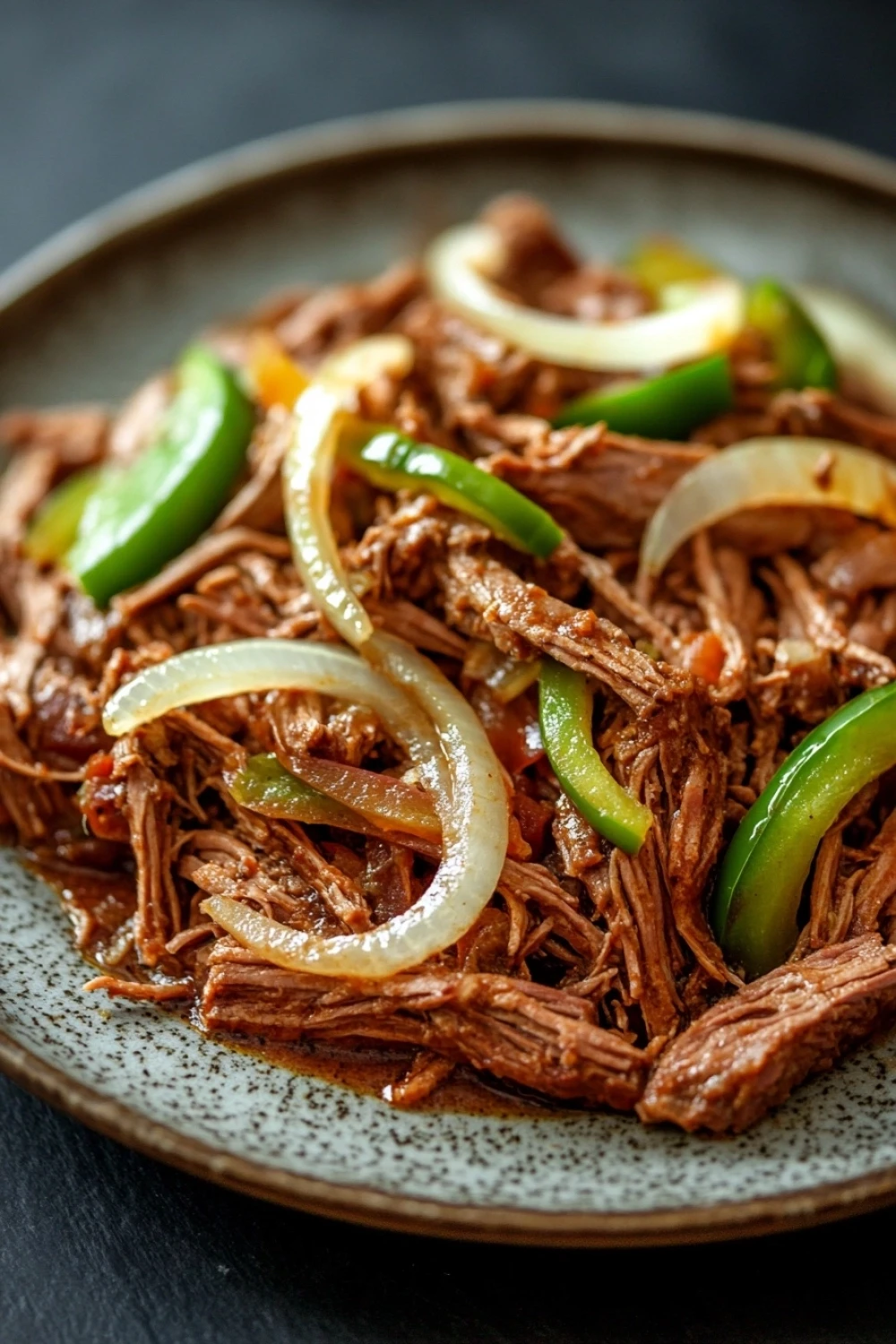 Italian beef slow cooker - the image is a close-up of a plate of food. the plate is round and has a speckled pattern. the food appears to be a dish made with shredded beef, sliced onions, and green bell peppers. the beef is cooked to a medium-rare and the onions are sliced into thin strips. the bell peppers are bright green and appear to be seasoned with spices. the dish is garnished with a sprinkle of herbs and spices, adding a pop of color to the plate. the background is dark, making the plate and its contents stand out.