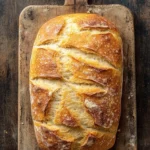 Kefir bread with yeast - the image shows a freshly baked loaf of bread on a wooden cutting board. the bread is golden brown in color and has a criss-cross pattern on the top. it appears to be freshly baked and is sitting on a dark wooden surface. the cutting board is made of light-colored wood and has two handles on either side. a string is tied around the top of the bread, possibly for hanging. the background is blurred, but it seems to be a rustic kitchen countertop.