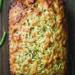 Keto zucchini sandwich bread - the image is a close-up of a freshly baked loaf of bread on a wooden surface. the bread appears to be golden brown in color and has a crumbly texture. on top of the bread, there are small pieces of zucchini and cheese scattered across the surface. there are also some green beans scattered around the loaf. the overall appearance is rustic and appetizing.