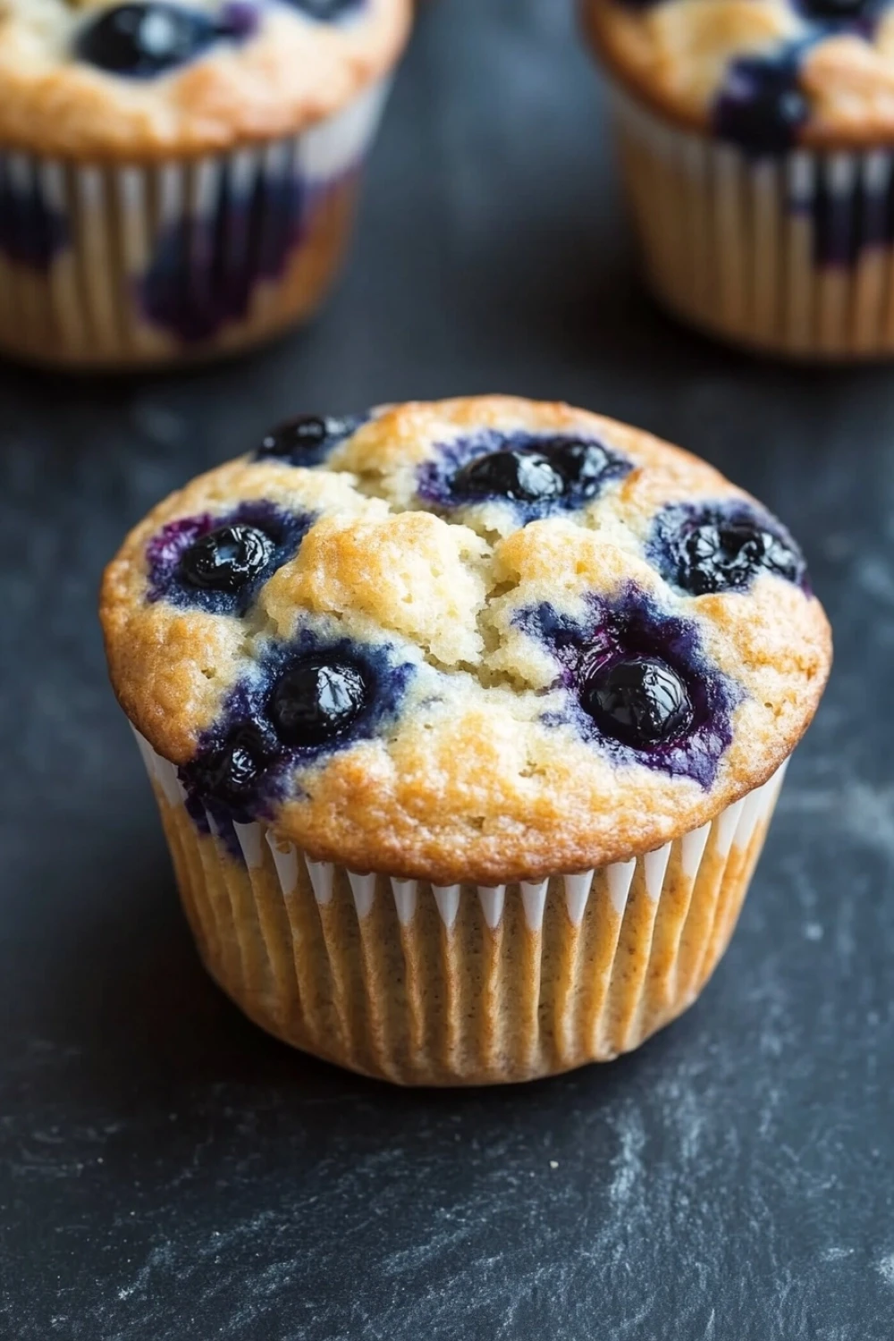 Kodiak muffin greek yogurt - the image shows three freshly baked blueberry muffins on a dark grey textured surface. the muffins are golden brown in color and have a crumbly texture. they are topped with fresh blueberries, which are visible in the center of each muffin. the blueberries are arranged in a circular pattern around the edges of the muffins, with some overlapping each other. the background is blurred, but it appears to be a kitchen countertop.