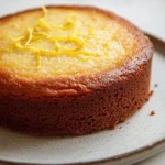 Lemon cake no milk - the image is a close-up of a round cake on a white plate. the cake appears to be a chocolate cake with a golden brown crust on top. the top of the cake is covered in a layer of yellow frosting, which is drizzled with a thin layer of orange zest. the frosting is smooth and shiny, and the cake looks freshly baked. the plate is sitting on a marble countertop.