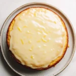 Lemon cake vintage - the image is a close-up of a round cake on a white marble countertop. the cake appears to be a lemon cake with a golden brown crust and a creamy white frosting on top. the frosting is drizzled over the top of the cake, creating a smooth and glossy texture. the plate it is sitting on is round and has a speckled pattern around the edges. the background is plain white, making the cake stand out.