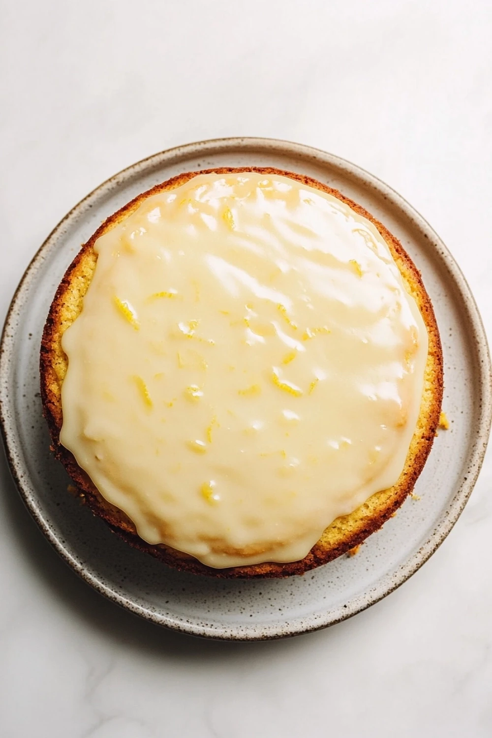 Lemon cake vintage - the image is a close-up of a round cake on a white marble countertop. the cake appears to be a lemon cake with a golden brown crust and a creamy white frosting on top. the frosting is drizzled over the top of the cake, creating a smooth and glossy texture. the plate it is sitting on is round and has a speckled pattern around the edges. the background is plain white, making the cake stand out.