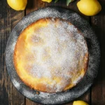 Lemon cake with italian ingredients - the image shows a round cake on a black plate with a dusting of powdered sugar on top. the cake appears to be freshly baked and has a golden brown crust. there are several lemons scattered around the plate, some of which are whole and some are still attached to the plate. the background is a wooden table with a few green leaves scattered around. the overall mood of the image is rustic and homey.