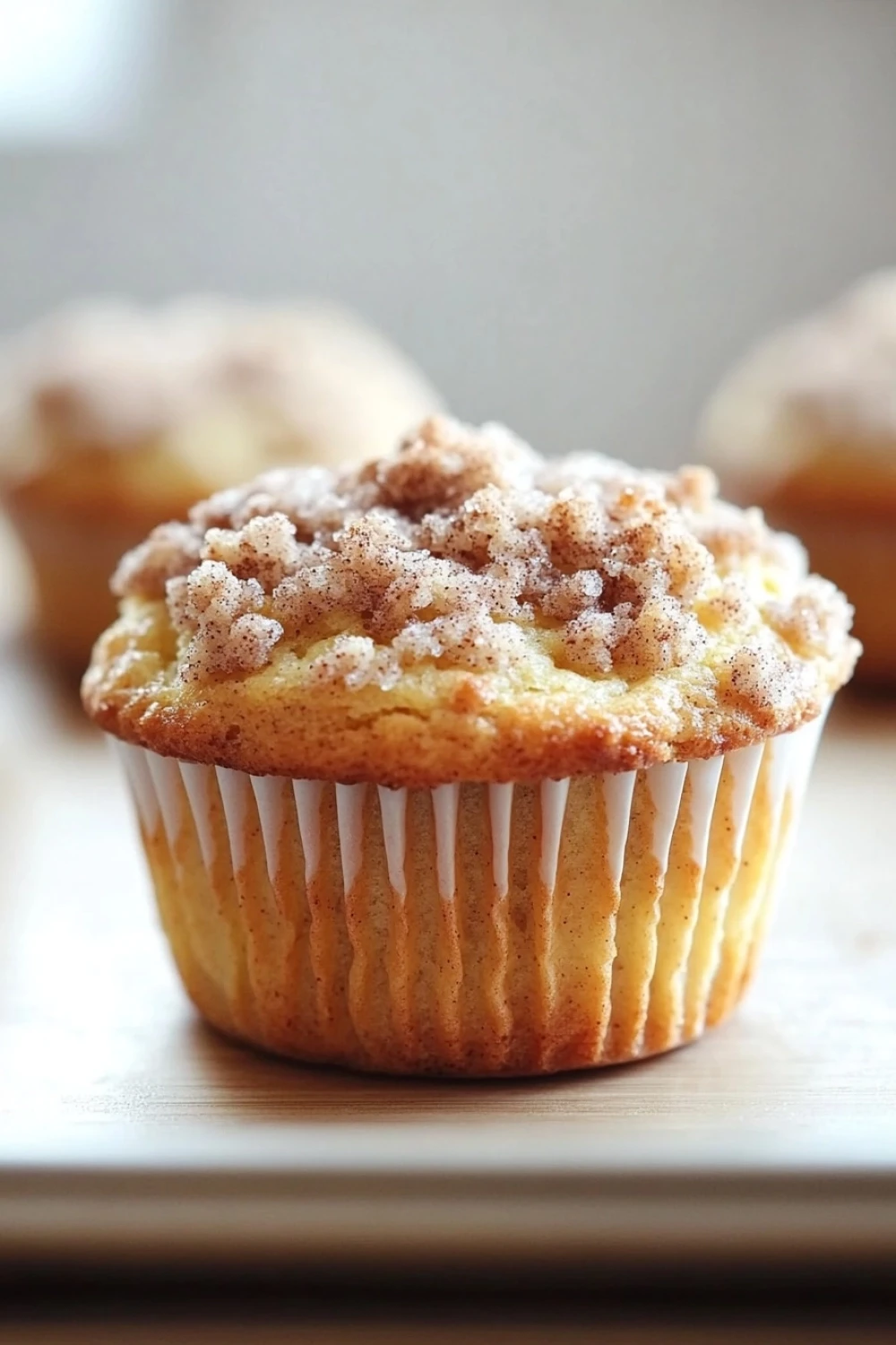 Lemon streusel muffin - the image shows a close-up of a freshly baked muffin on a white plate. the muffin is golden brown in color and has a crumbly texture on top. it is covered in a light dusting of powdered sugar, giving it a shiny appearance. in the background, there are other muffins visible, but they are slightly blurred. the plate is on a wooden table, and the background is out of focus.