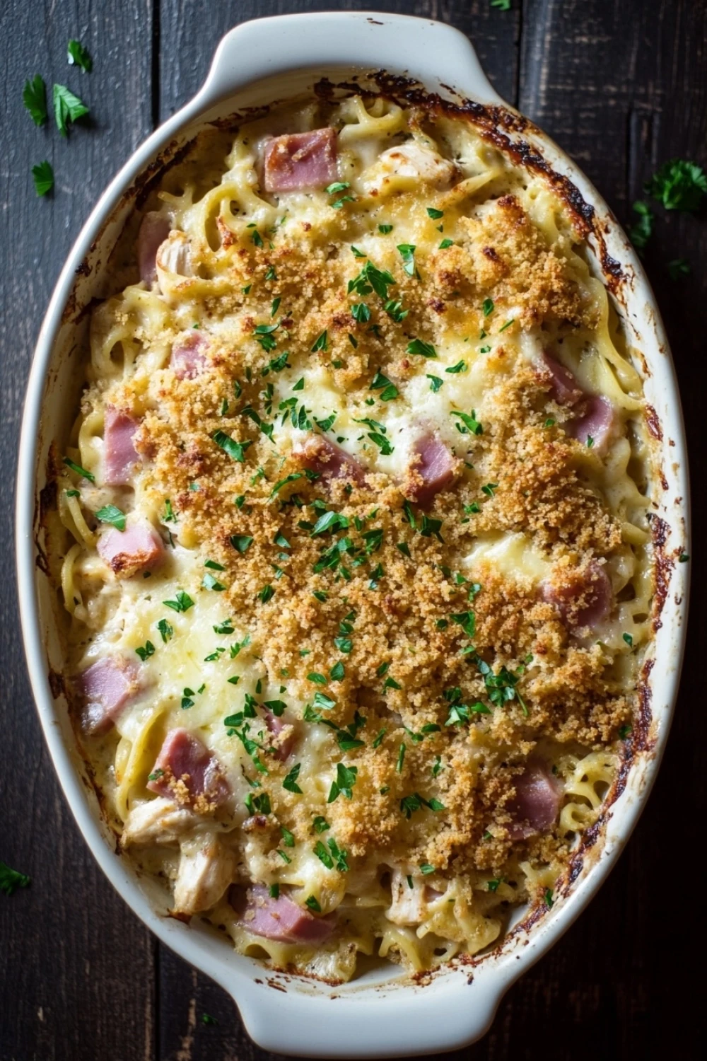 Make ahead chicken cordon bleu casserole - the image shows a white ceramic casserole dish filled with a dish of macaroni and cheese. the dish appears to be freshly baked and has a golden brown crust on top. the cheese is melted and bubbly, and there are chunks of ham scattered throughout the dish. there are also a few sprigs of parsley scattered around the dish, adding a pop of green color. the background is a dark wooden table, and the overall mood of the image is warm and inviting.