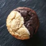 Muffin bicolore allo yogurt - the image is a close-up of a muffin on a black textured surface. the muffin appears to be freshly baked and has a golden brown color. it has a crumbly texture and is filled with a creamy white filling. the filling is oozing out of the top of the muffin, revealing the soft, fluffy interior. the background is blurred, making the muffins the focal point of the image.