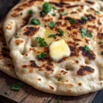 Nann bread no yogurt - the image shows a flatbread on a wooden cutting board. the flatbread is golden brown in color and appears to be freshly baked. on top of the flatbread, there is a small amount of melted butter and a sprig of fresh parsley. the cutting board is placed on a dark wooden table. the background is blurred, but it seems to be a kitchen countertop.