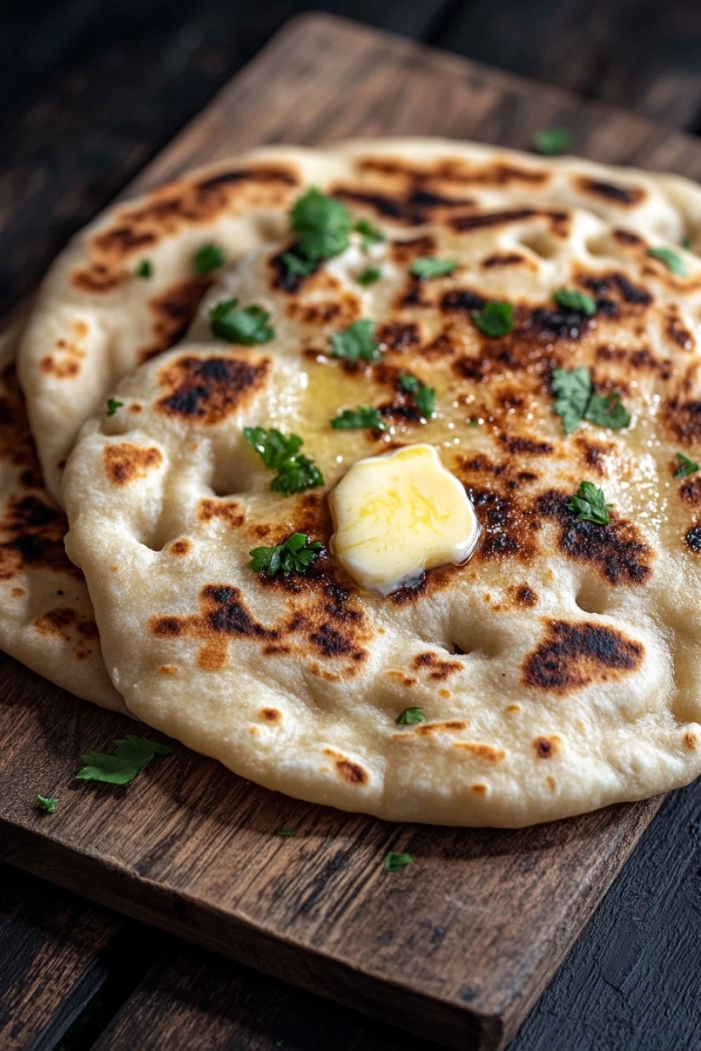 Nann bread no yogurt - the image shows a flatbread on a wooden cutting board. the flatbread is golden brown in color and appears to be freshly baked. on top of the flatbread, there is a small amount of melted butter and a sprig of fresh parsley. the cutting board is placed on a dark wooden table. the background is blurred, but it seems to be a kitchen countertop.