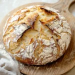 No knead 5 minute artisan bread - the image shows a round loaf of bread on a wooden cutting board. the bread appears to be freshly baked and has a golden brown crust on top. the edges of the bread are slightly frayed, indicating that it has been freshly baked. the cutting board is placed on a white countertop with a white cloth napkin next to it. the background is blurred, but it seems to be a kitchen countertop.