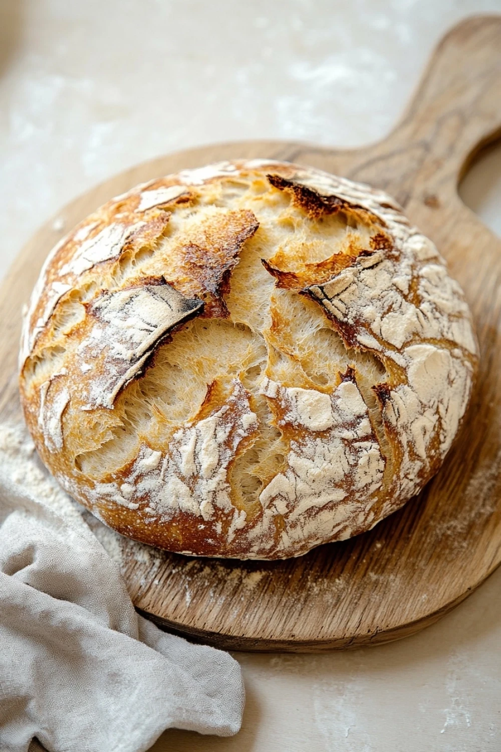 No knead 5 minute artisan bread - the image shows a round loaf of bread on a wooden cutting board. the bread appears to be freshly baked and has a golden brown crust on top. the edges of the bread are slightly frayed, indicating that it has been freshly baked. the cutting board is placed on a white countertop with a white cloth napkin next to it. the background is blurred, but it seems to be a kitchen countertop.
