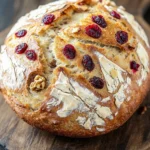 No knead bread cranberry walnut - the image is a close-up of a freshly baked loaf of bread on a wooden cutting board. the bread is golden brown in color and has a crumbly texture. it is topped with sliced cranberries and walnuts, giving it a crunchy appearance. the cranberries are bright red and appear to be ripe and juicy. the walnuts are scattered throughout the bread, adding a nutty flavor to the dish. the cutting board is made of dark wood and is placed on a rustic wooden table.