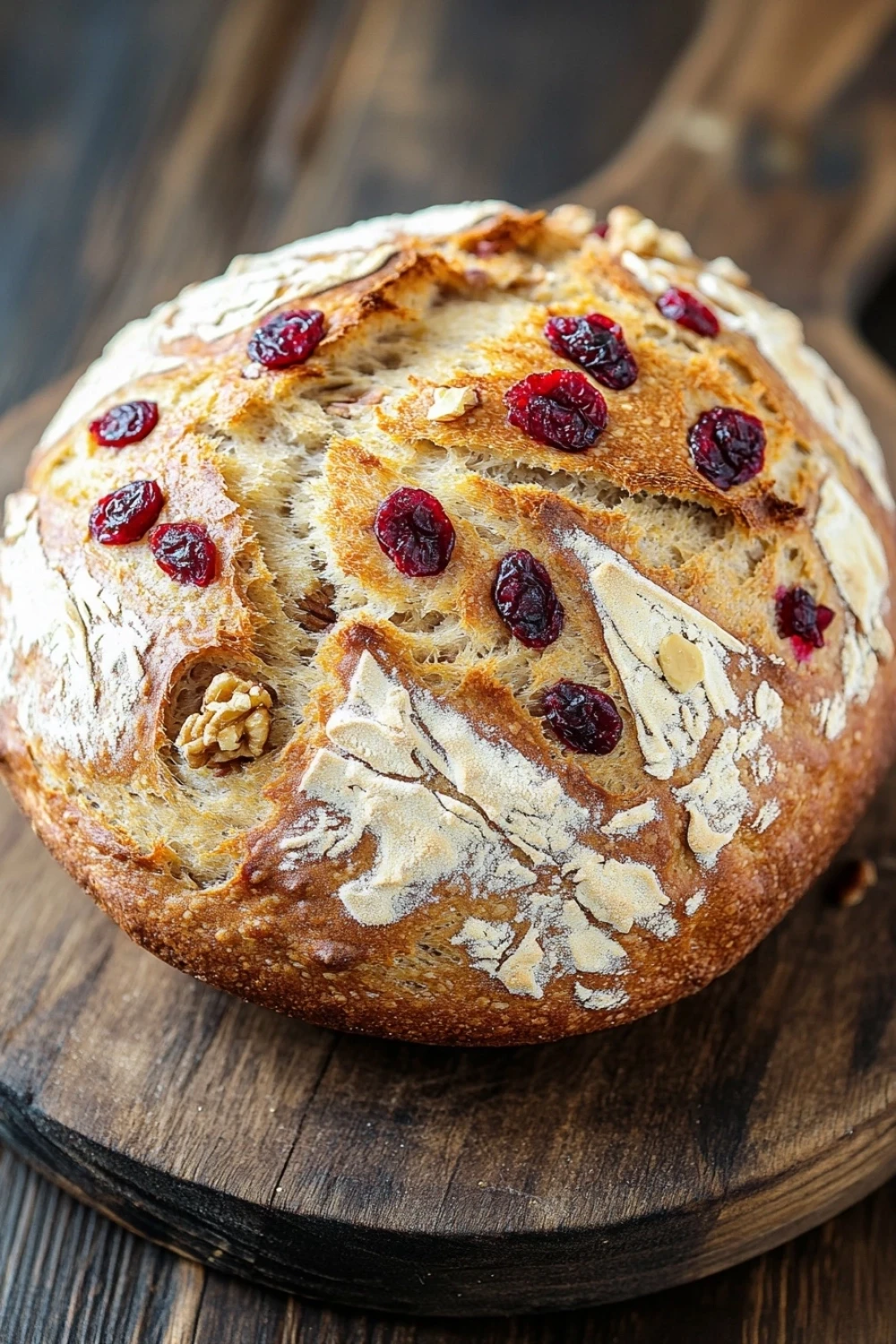 No knead bread cranberry walnut - the image is a close-up of a freshly baked loaf of bread on a wooden cutting board. the bread is golden brown in color and has a crumbly texture. it is topped with sliced cranberries and walnuts, giving it a crunchy appearance. the cranberries are bright red and appear to be ripe and juicy. the walnuts are scattered throughout the bread, adding a nutty flavor to the dish. the cutting board is made of dark wood and is placed on a rustic wooden table.
