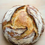 No knead bread dutch oven rise - the image is a close-up of a round loaf of bread on a wooden surface. the bread appears to be freshly baked and has a golden brown crust on top. the inside of the bread is visible, with a swirl pattern of white and brown swirls. the edges of the crust are slightly frayed, indicating that the bread has been freshly baked. the background is a light-colored wooden surface, which contrasts with the warm tones of the wood. the overall mood of the image is rustic and cozy.