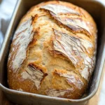 No knead sourdough bread loaf pan - the image shows a loaf of bread in a metal loaf pan on a wooden cutting board. the bread appears to be freshly baked, with a golden brown crust and a soft, fluffy interior. the loaf is sitting on a white countertop with a white cloth napkin on the side. the background is blurred, but it seems to be a kitchen countertop.