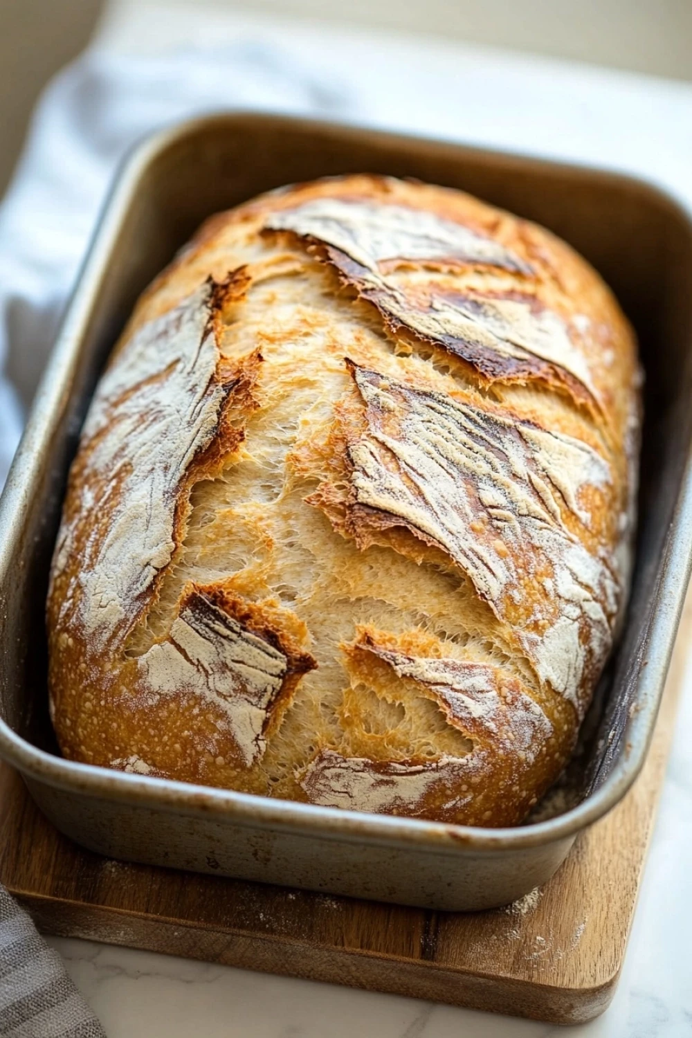 No knead sourdough bread loaf pan - the image shows a loaf of bread in a metal loaf pan on a wooden cutting board. the bread appears to be freshly baked, with a golden brown crust and a soft, fluffy interior. the loaf is sitting on a white countertop with a white cloth napkin on the side. the background is blurred, but it seems to be a kitchen countertop.
