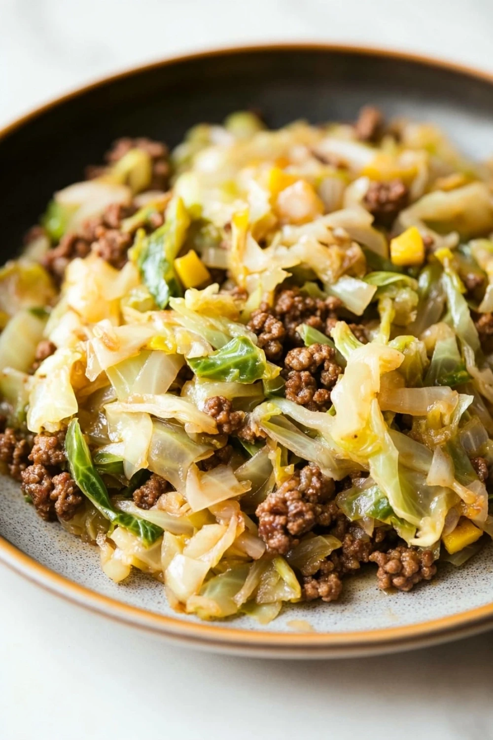 One pan beef cabbage skillet - the image is a close-up of a dish in a black bowl. the dish appears to be a stir-fry with cabbage, ground beef, and other vegetables. the cabbage is cut into thin strips and is mixed with diced yellow and green bell peppers. the ground beef is dark brown and looks crispy on the outside. the vegetables are bright green and look fresh and vibrant. the bowl is sitting on a white countertop.
