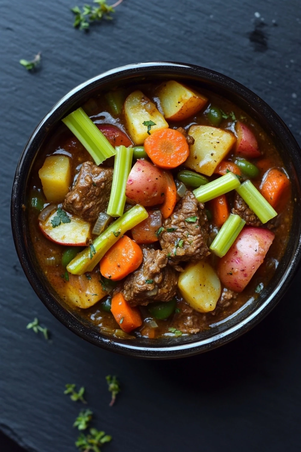 One pot beef and veggies - the image is a close-up of a bowl of beef stew. the stew is made with chunks of beef, carrots, potatoes, and celery. the beef is cooked in a rich, dark brown broth and is garnished with chopped herbs. the vegetables are cut into small cubes and are arranged in a circular pattern around the beef. the bowl is black and is sitting on a dark, textured surface. there are a few sprigs of parsley scattered around the bowl.