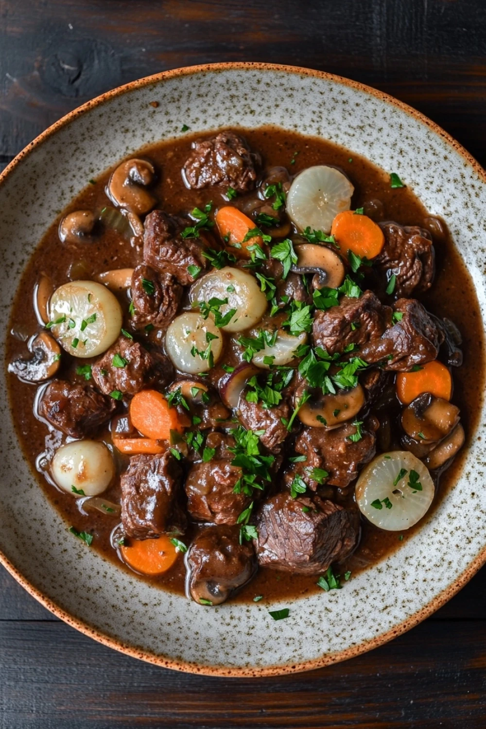 One pot beef bourguignon - the image is a close-up of a bowl of beef stew. the stew is made with chunks of beef, mushrooms, and carrots, all cooked in a rich, dark brown sauce. the beef is cooked to a medium-rare and appears to be tender and juicy. the mushrooms are sliced and arranged in a circular pattern around the beef. the carrots are bright orange and are scattered throughout the stew, adding a pop of color to the dish. there are also small pieces of lemon wedges and parsley scattered throughout. the bowl is sitting on a wooden table, and the background is blurred, making the stew the focal point of the image.