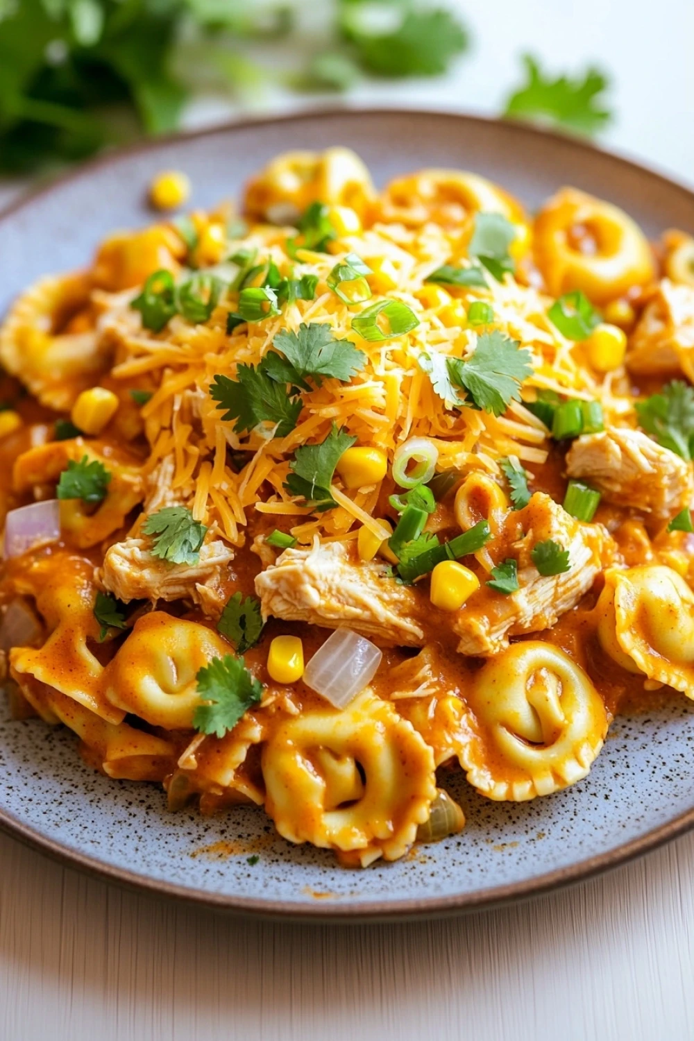 One pot chicken enchilada tortellini - the image is a close-up of a plate of pasta dish. the pasta appears to be fusilli, which is a type of spiral-shaped pasta, and is covered in a creamy orange sauce. the sauce is drizzled over the top of the pasta, with chunks of chicken and corn visible. there are also chunks of red onion and cilantro scattered throughout the dish, adding a pop of green color. the plate is blue with a speckled pattern, and there are a few sprigs of cilantro on the side. the background is a white tablecloth, and the overall color scheme of the dish is warm and inviting.