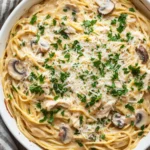 One pot chicken tetrazzini - the image shows a large white casserole dish filled with spaghetti and mushrooms. the spaghetti is cooked al dente and is covered in a creamy sauce with chunks of chicken and parmesan cheese. the dish is garnished with fresh parsley. the background is a wooden table with a gray and white striped napkin on the side. the overall color scheme of the dish is warm and inviting.