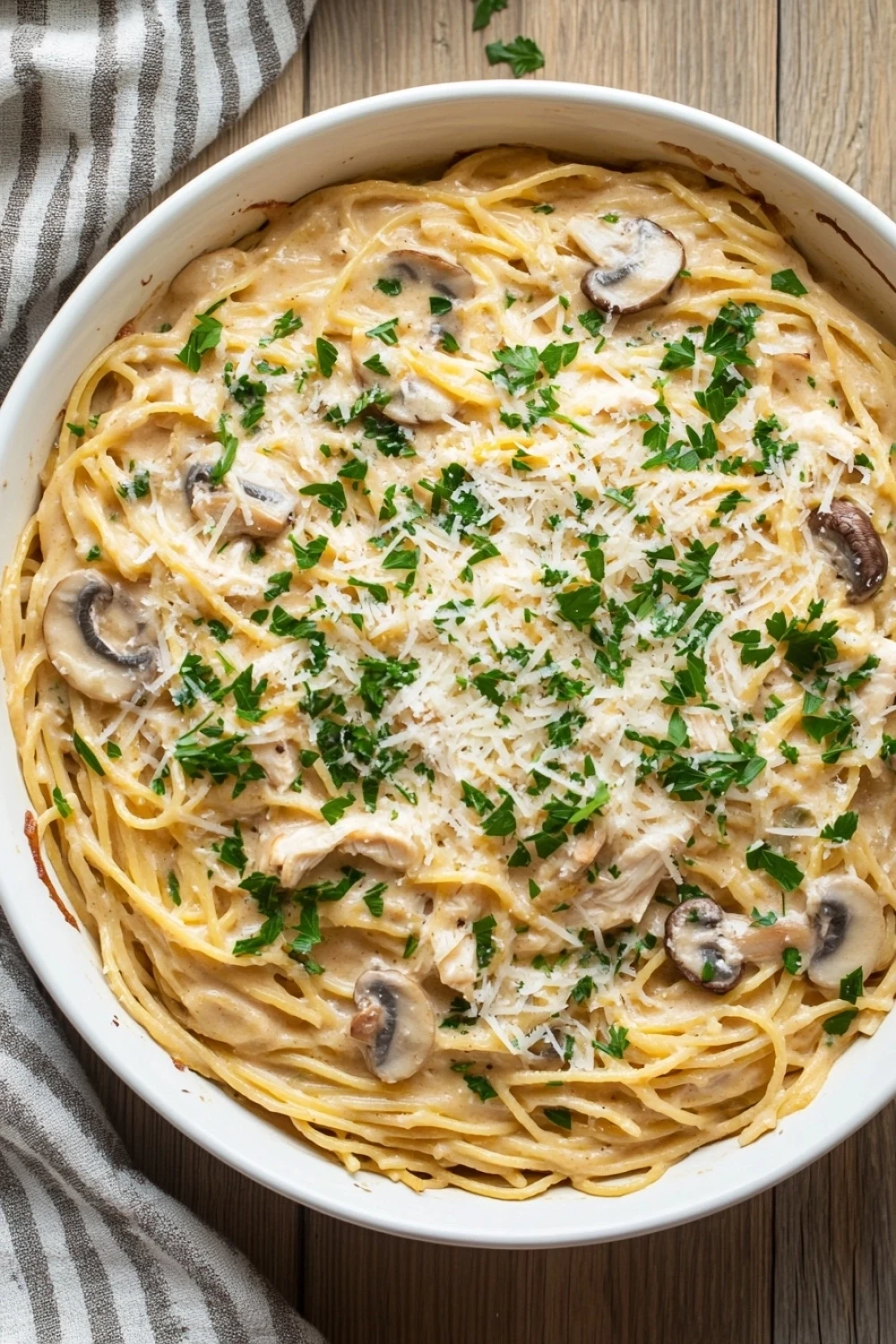 One pot chicken tetrazzini - the image shows a large white casserole dish filled with spaghetti and mushrooms. the spaghetti is cooked al dente and is covered in a creamy sauce with chunks of chicken and parmesan cheese. the dish is garnished with fresh parsley. the background is a wooden table with a gray and white striped napkin on the side. the overall color scheme of the dish is warm and inviting.