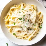 One pot creamy fettuccine - the image shows a bowl of tagliatelle pasta on a white marble countertop. the pasta is cooked al dente and is topped with grated parmesan cheese and fresh parsley. the bowl is white and there is a gray cloth napkin on the right side of the image. the background is blurred, but it appears to be a kitchen countertop with a few scattered parsley leaves.