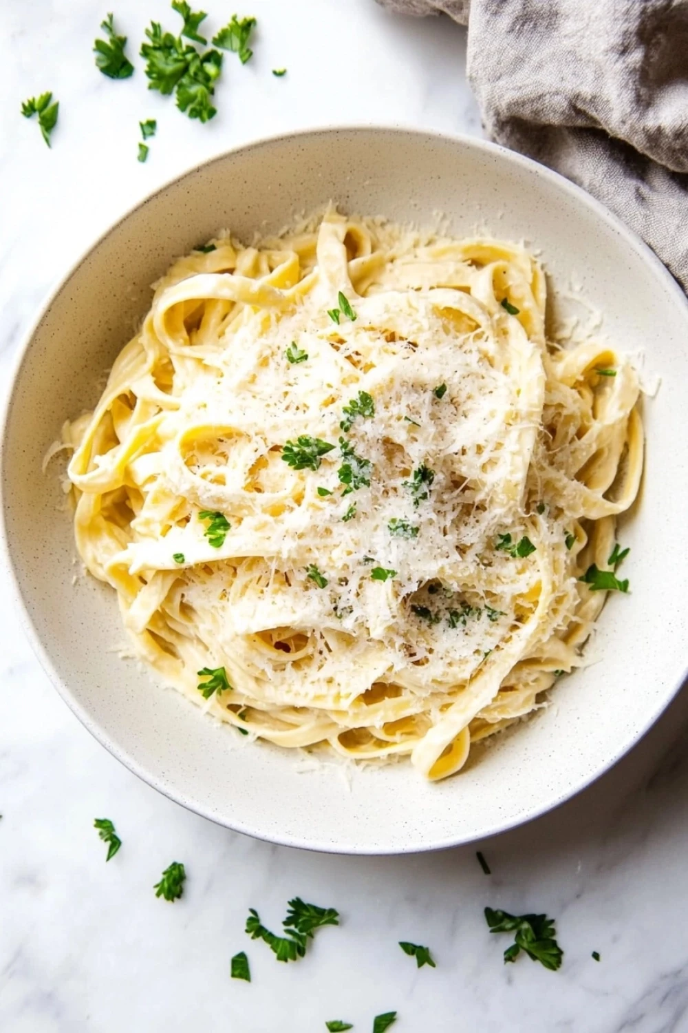 One pot creamy fettuccine - the image shows a bowl of tagliatelle pasta on a white marble countertop. the pasta is cooked al dente and is topped with grated parmesan cheese and fresh parsley. the bowl is white and there is a gray cloth napkin on the right side of the image. the background is blurred, but it appears to be a kitchen countertop with a few scattered parsley leaves.